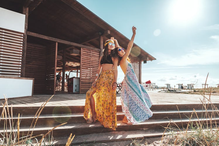 Two female friends wearing sunglasses and maxi skirts smile and pose on the steps of a beach shack o...