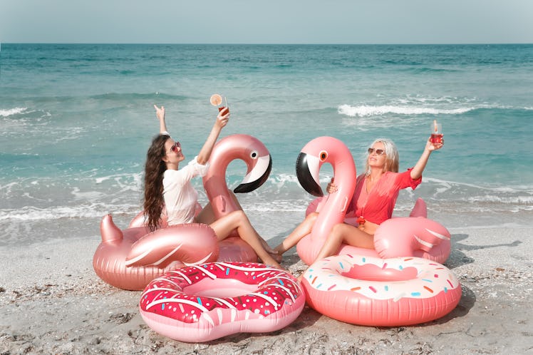 Two women enjoy the beach on a sunny day while sitting on pink flamingo pool floats and holding drin...