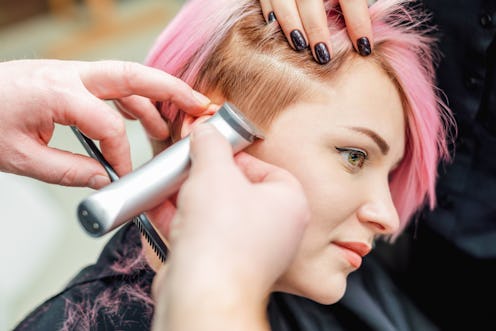 a barber shaves temple woman pink hair with electric shaver in beauty salon, close up hairdresser sh...