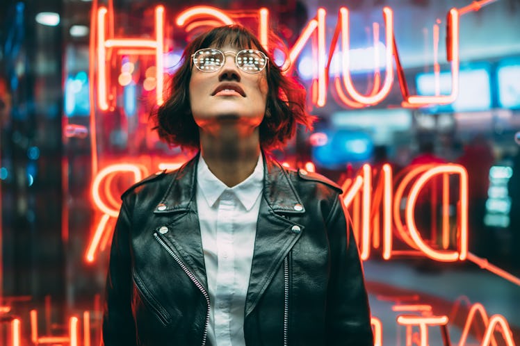 Stylish brunette woman in trendy apparel and eyewear looking up enjoying nightlife in city. Gorgeous...