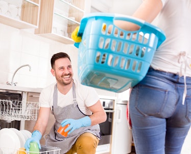 Young couple doing chores together. Man loading dishwasher and wife holding laundry basket