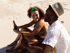 A happy couple chats and holds their beer bottles while sitting on a beach on a sunny day.