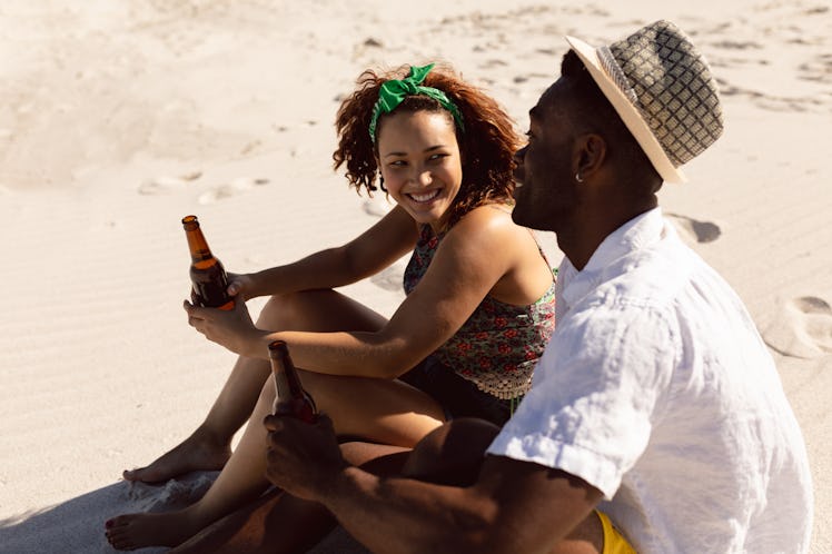 A happy couple chats and holds their beer bottles while sitting on a beach on a sunny day.