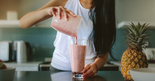 Woman pouring healthy smoothie in glass from grinder jar on kitchen counter. Female preparing fresh ...