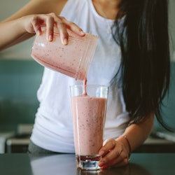 Woman pouring healthy smoothie in glass from grinder jar on kitchen counter. Female preparing fresh ...