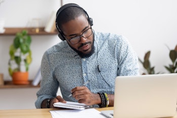 Focused african business man in headphones writing notes in notebook watching webinar video course, ...
