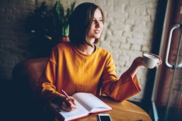 A woman in a yellow sweater writes in a sketchbook in a café.