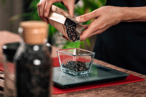 Weighing coffee grains on digital scale. Hands of female barista pouring roasted coffee grains on a ...