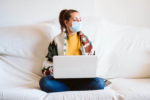 young woman working on laptop at home, sitting on the couch, wearing protective mask. Stay home conc...