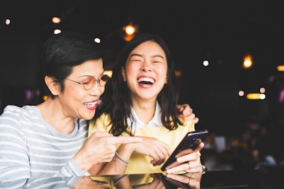 An asian mother and daughter laugh and smile while looking at a cell phone in a restaurant.