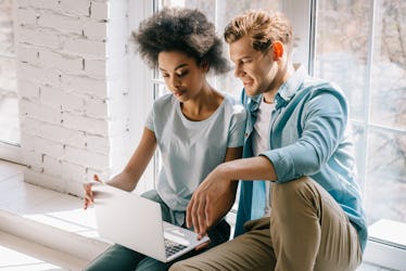 A couple sits on a windowsill in front of a big white window while looking at a laptop.