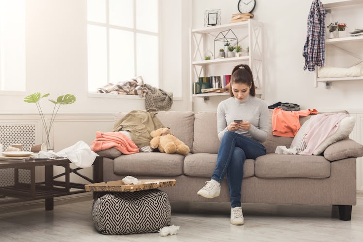 Desperate helpless woman sitting on sofa in messy living room. and chatting on mobile, surrounded by...