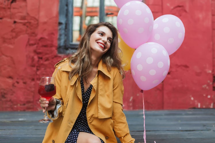 A woman wearing a yellow jacket, holds a glass of sangria and some pink balloons, while sitting outs...