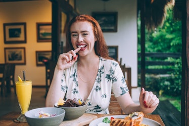 A happy redhead woman smiles at a table with a mimosa, chips, and sandwich.