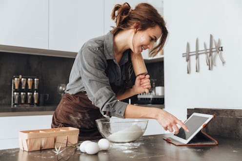 Image of young pretty lady standing in kitchen and cooking the dough. Looking at tablet computer.