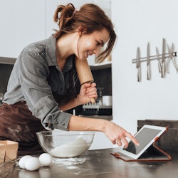 Image of young pretty lady standing in kitchen and cooking the dough. Looking at tablet computer.
