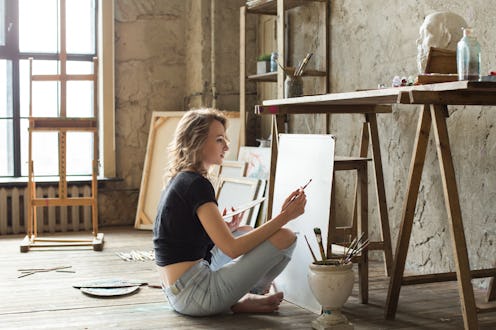 Woman painter sitting on the floor in front of the canvas and drawing. Artist studio interior. Drawi...
