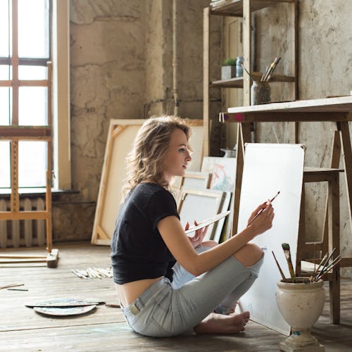 Woman painter sitting on the floor in front of the canvas and drawing. Artist studio interior. Drawi...