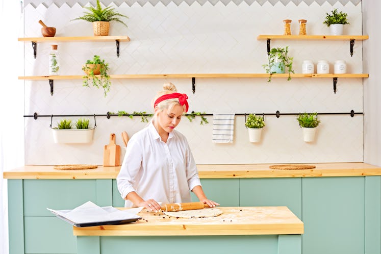A blonde woman with a red headband and white button-down shirt rolls out dough in her bright kitchen...