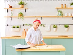 A blonde woman with a red headband and white button-down shirt rolls out dough in her bright kitchen...