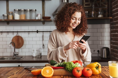 A happy woman looks at her phone, while standing in her kitchen with vegetables in front of her. 