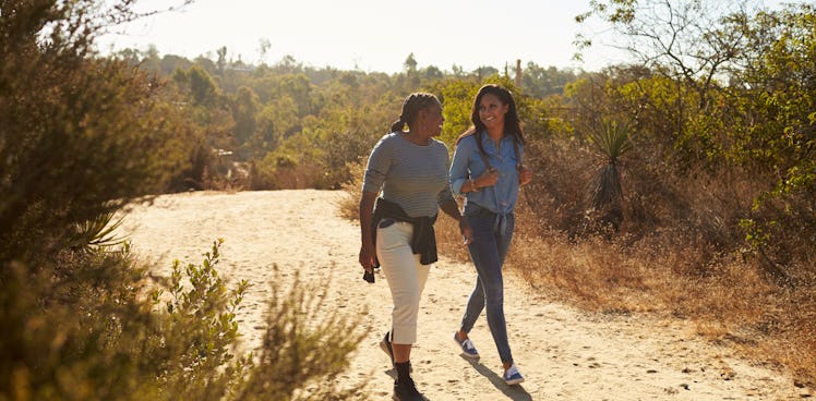 A mother and daughter chat while hiking a dirt trail in the sunshine.