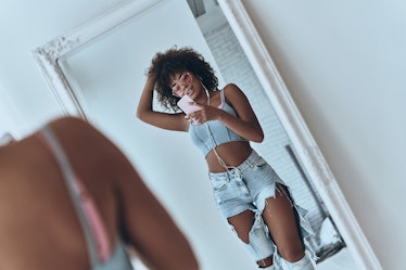 A woman wearing ripped jeans, a crop top, and pink sunglasses takes a mirror selfie in her room.