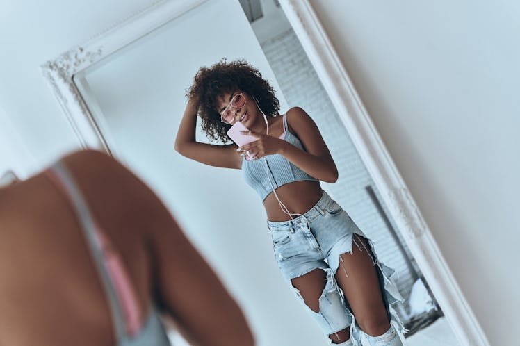 A woman wearing ripped jeans, a crop top, and pink sunglasses takes a mirror selfie in her room.