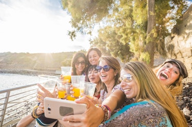 A group of women hold up cups of orange juice while snapping a selfie by the water on a sunny day.