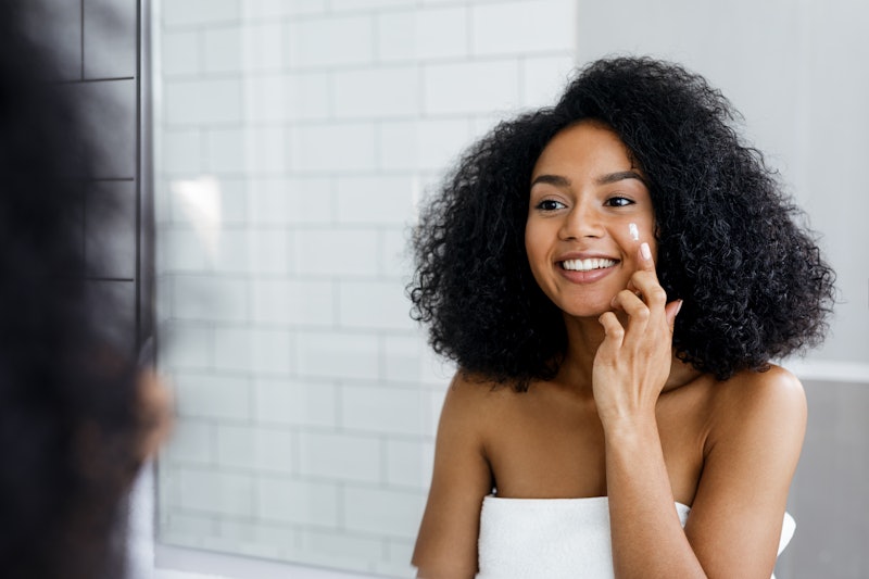 Happy young woman applying face cream with finger in bathroom