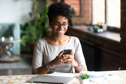 Cheerful african student black woman sitting at table studying using laptop reading a book, take a b...