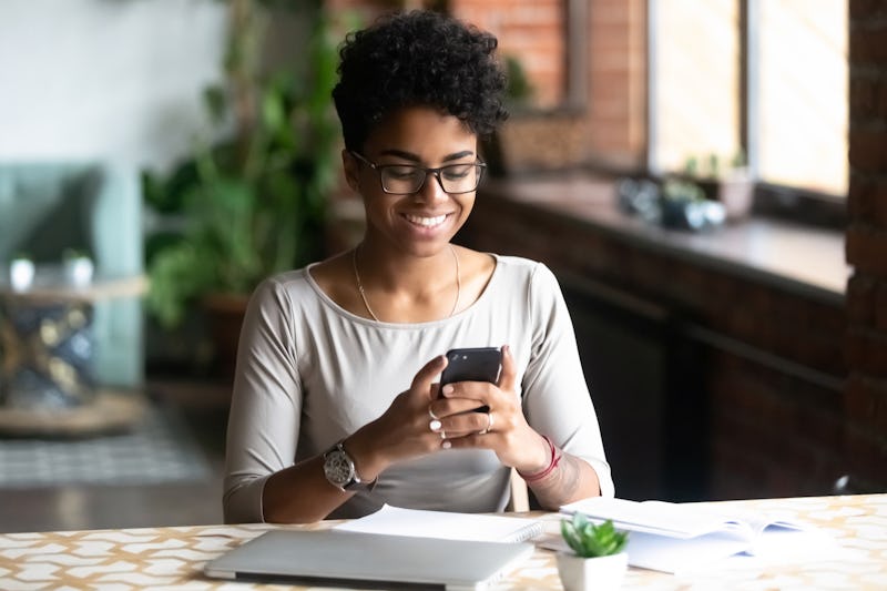 Cheerful african student black woman sitting at table studying using laptop reading a book, take a b...