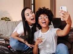 Two roommates smile for a selfie, while holding wine glasses on the couch. 