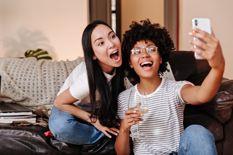 Two roommates smile for a selfie, while holding wine glasses on the couch.