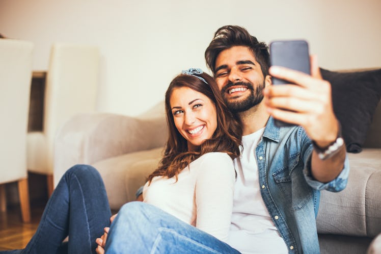 A happy couple sits on the floor at home while taking a selfie on their phone. 
