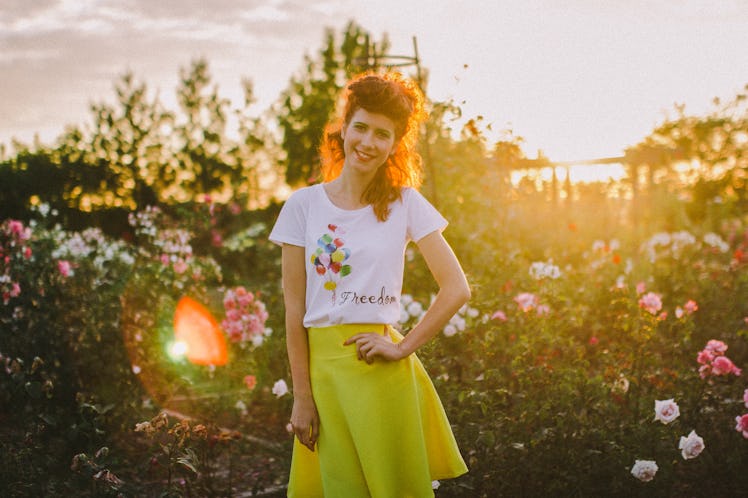 A redhead woman wears a yellow skirt and balloon shirt outside, next to some rose bushes. 