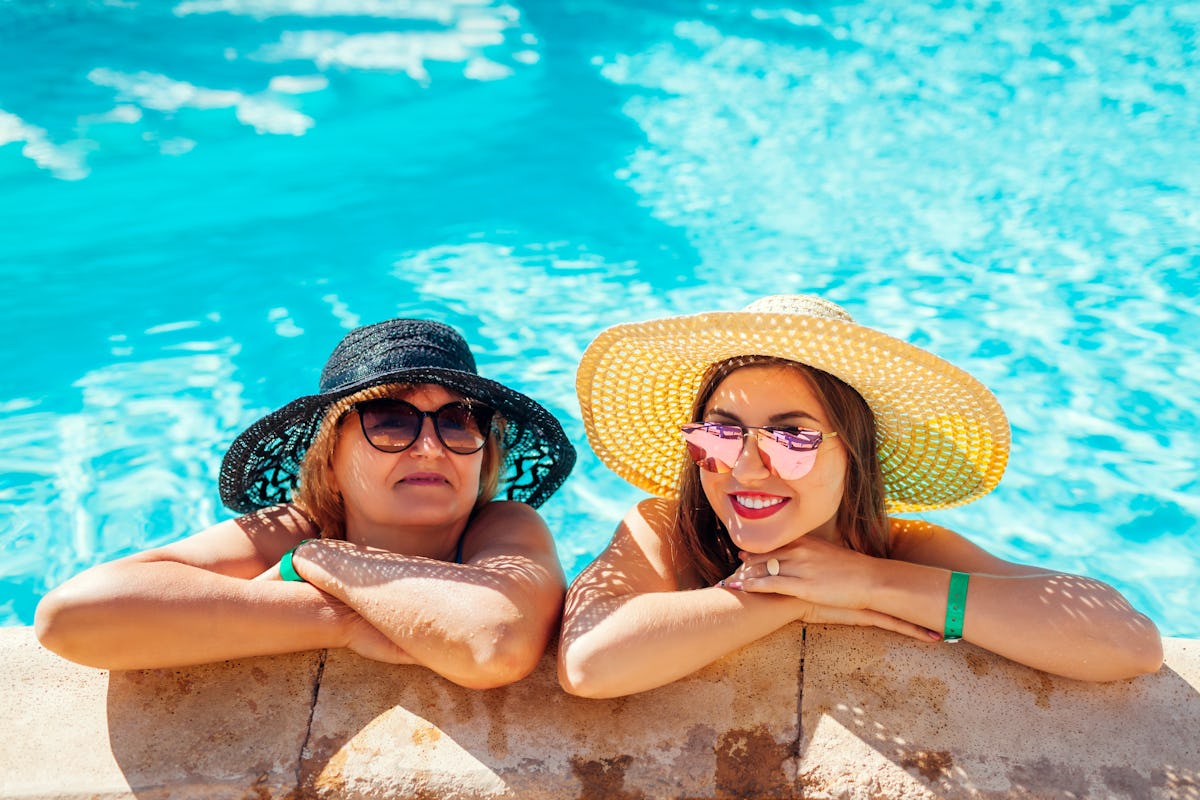 A mom and adult daughter smile and relax in a pool on a sunny day, while wearing sunglasses and sun hats.
