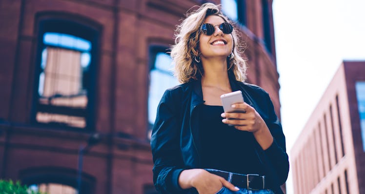 A happy woman, wearing sunglasses, stands with her phone in her hand outside. 