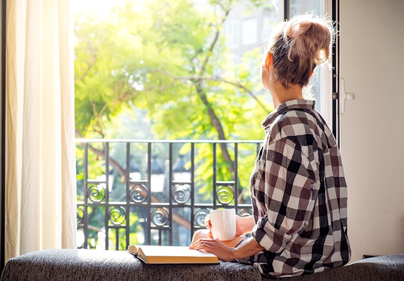 Young pretty woman  sitting at opened window drinking coffee and looking outside enjoys of rest