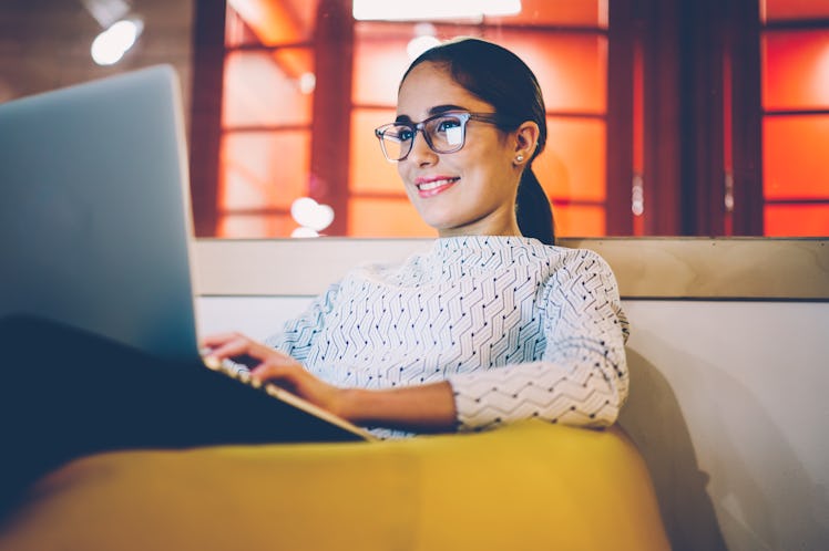 Smiling woman in eyewear watching video on laptop computer working on freelance in cozy coworking in...