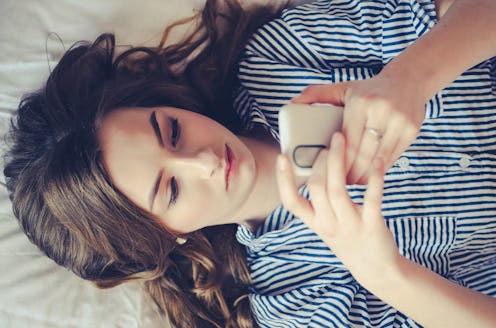 Closeup of sad teenage girl lying in bed using her mobile. Young pretty woman with bored expression ...