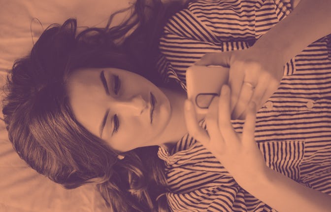 Closeup of sad teenage girl lying in bed using her mobile. Young pretty woman with bored expression ...