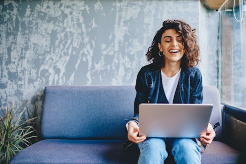 A happy woman sits on her couch at home with her laptop computer. 