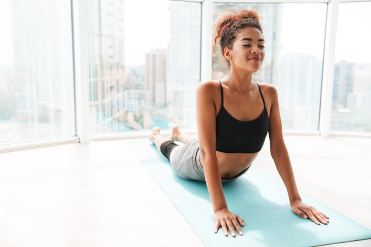 A woman smiles while stretching on her yoga mat in her bright apartment.