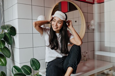 A woman in her pajamas sits on her bathroom counter, holding onto an eye mask on her head. 