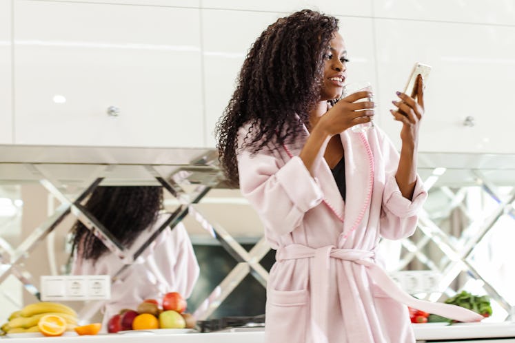 A woman in her pink robe stands in her kitchen, holding her phone and a glass of water.