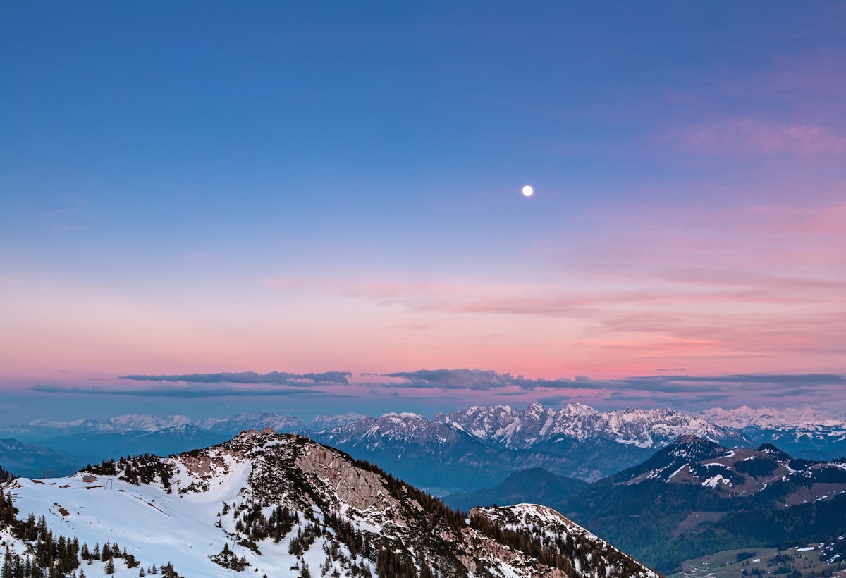 A full moon is above snow-capped mountains and a pink sky after sunset.