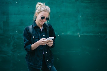 Smiling caucasian young woman sending message on smartphone standing near promotional copy space bac...