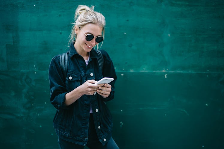 A happy woman looks down at phone while texting in front of green wall outside.