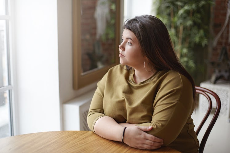 Loneliness concept. Young brunette plus size woman with black hair sitting at cafe table, feeling lo...